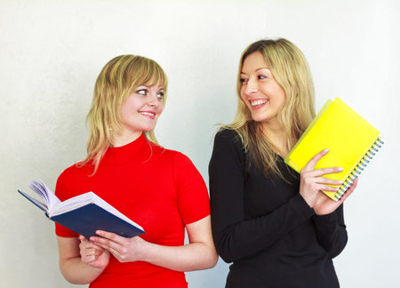 two beautiful young girl with long blond hair are holding a book and notebook, look at each other,...