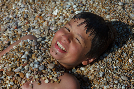 happy laughing boy of twelve, sprinkled with pebbles on the beachの写真素材