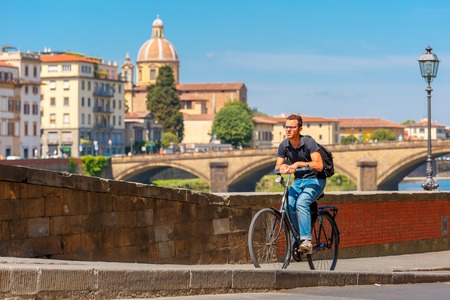 Florence, Italy - May 19, 2015: Young Caucasian man cyclist with glasses on the quay of river Arno, San Frediano in Cestello and Ponte alla Carraia on the background, Tuscany.のeditorial素材