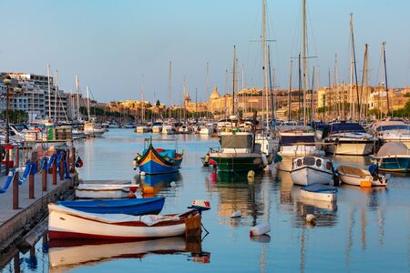 Valletta harbour with yachts and multicolored fishing boats Luzzu with eyes, church and fortress, illuminated by sunset light, Maltaの写真素材