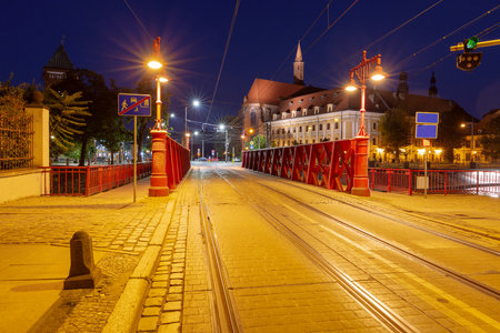 Night view of the Red Bridge in Wroclaw, Poland, illuminated by streetlights, with tram tracks and historical buildings in the backgroundの写真素材