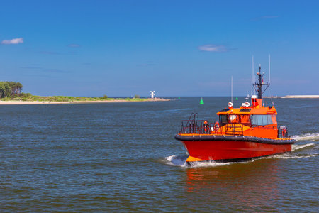 An orange pilot boat navigating the mouth of the Swina River in Swinoujscie, Poland, with the Stawa Mlyny beacon visible on the breakwater in the backgroundの写真素材