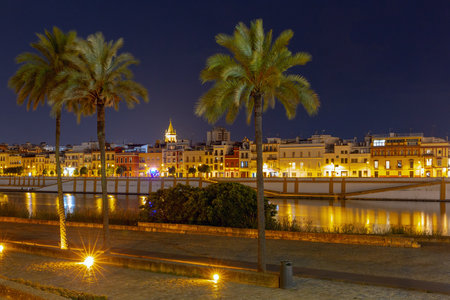 Nighttime view of the illuminated Triana district in Seville, Spain, showcasing its traditional colorful buildings along the Guadalquivir Riverの写真素材