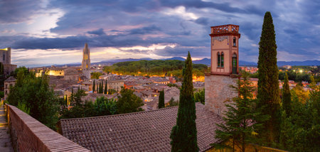 A panoramic view of Girona, Spain, at dusk, featuring the Church of Sant Feliu, historic rooftops, and a tower with decorative detailsの写真素材