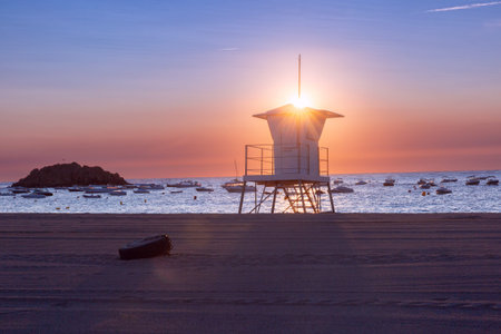 Silhouetted lifeguard tower and boats at sunrise on the beach in Tossa de Mar, Spainの写真素材