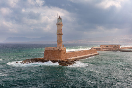 Historic lighthouse on the sea wall at the harbor of Chania in Crete, Greece with waves and cloudy skyの写真素材