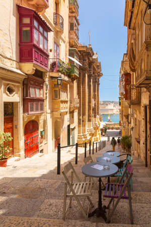 Colorful traditional wooden balconies on a narrow stone staircase street in Valletta Malta with empty cafe tables in the foregroundの写真素材
