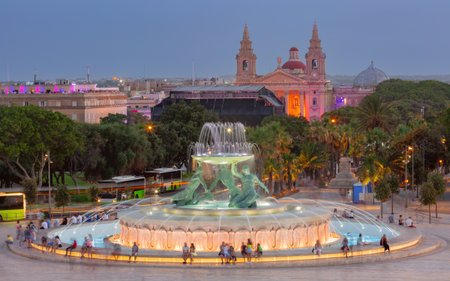 Evening view of illuminated Triton Fountain and Saint Publius Church in Valletta Malta with people in the squareの写真素材