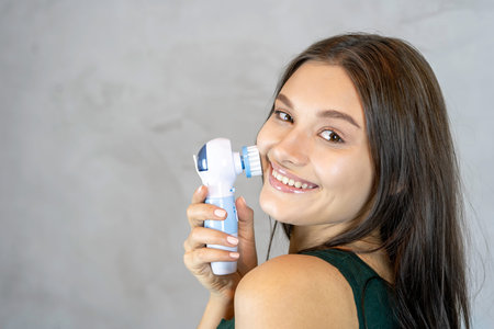 Young pretty woman doing skin care procedure. Cleaning face with machine on grey background.