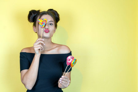 Beautiful happy smiling woman with lollipop and fashion hairstyle on yellow background. Copy space