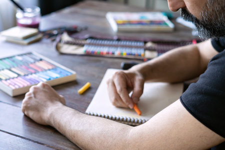 man painting with pencil and pastel crayon chalks on a white sheet of paper. box of pastel chalks and color pencils on the wooden tableの写真素材