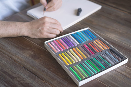 person painting with a pastel crayon chalk on a white sheet of paper with pallette of pastel chalks on the wooden table.の写真素材