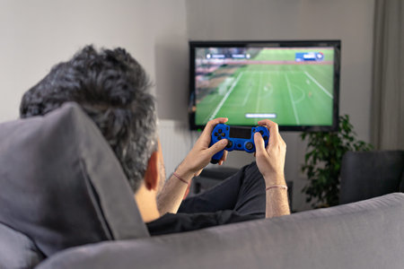 man playing video game at home sitting on coach sofa in front of television with play game concole...