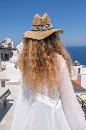 Beautiful young woman in white dress straw hat on white terrace balcony of house or hotel with Sea Viewの写真素材