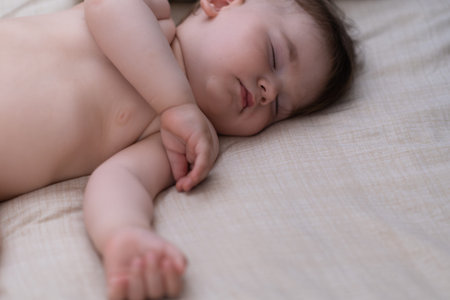 Sweet chubby little boy with closed eyes sleeping on comfortable bed spreading hands on white sheet...