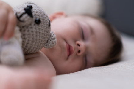Chubby baby boy with closed eyes sleeping in crib on soft white sheet hugging knitted bear adorable...