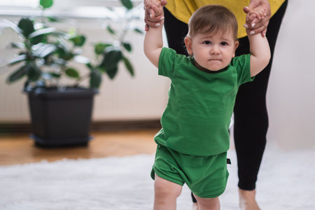 Chubby baby boy in green outfit learning to walk holding mother hands caring woman developing little son helping to make first steps on carpet in living room at homeの写真素材