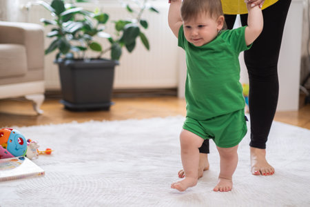 Loving mom helping chubby little son developing new skills at home cute baby boy in green outfit learning to walk on white fluffy rug holding hands of motherの写真素材