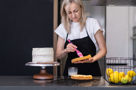 Focused blonde confectioner spreading white cream on cake layers with cooking brush standing at kitchen table woman in black apron making creamy pastry from organic products at homeの写真素材