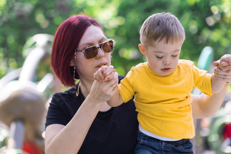 Caring mother in sunglasses holding hands of little son with Down syndrome helping to climb structure woman spending time with boy at kids playground on sunny weekendの写真素材