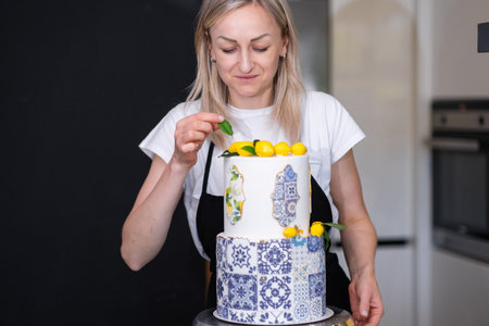 Satisfied housewife putting decorative green leaf near yellow lemons on top of two-tiered cake standing in kitchen small business of organic sugar free confectionery production at homeの写真素材