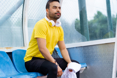 Bearded man with towel and headphones sitting on plastic chair in arbor at city sports stadium sportsman resting and team game on outdoor athletic arenaの写真素材