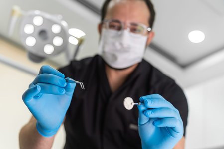 Male dentist in black uniform and gloves reaching to patient with mirror and tongs examining oral...
