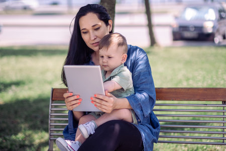 Young mother showing development game on tablet computer to cute baby son sitting together on bench in green urban park working online woman with little childの写真素材