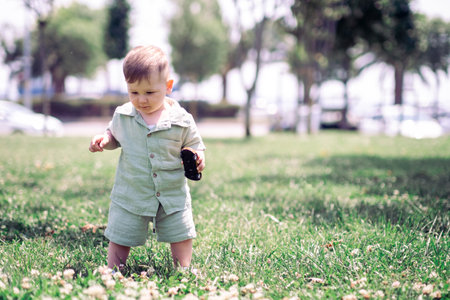 Cute little boy in cotton suit holding toy car standing on green lawn with flowers in summer urban park active infant child playing on meadow on sunny dayの写真素材