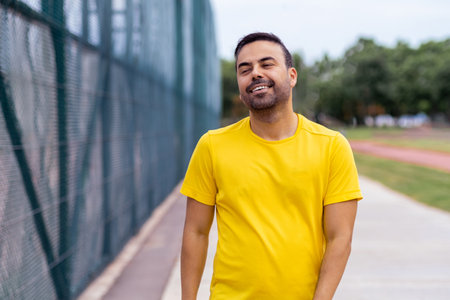 Smiling man in yellow t-shirt walking along road next to net fence on public sports ground in spring park positive young athlete exercising for wellbeingの写真素材