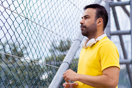 Man with earphones on neck and water bottle standing by metal braided fence on street walker spending time in urban park on spring day lifestyle and leisureの写真素材