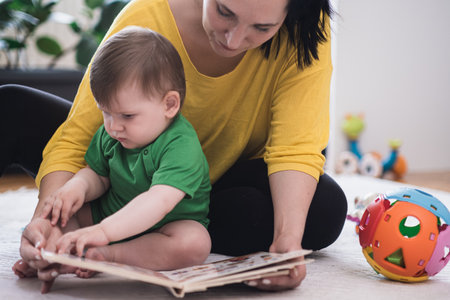 Chubby baby boy examining book with colorful pictures sitting near mother on white rug near toys woman educating little son at home developing new skillsの写真素材