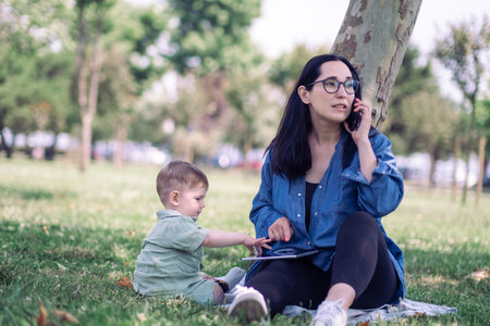 Infant child touching tablet screen while mother talking on mobile phone and sitting in tree shadow on urban garden grass lawn freelancer with playful son in parkの写真素材