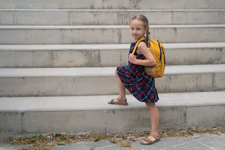 With a yellow backpack and a radiant smile, a cheerful blonde schoolgirl in her dress heads to...