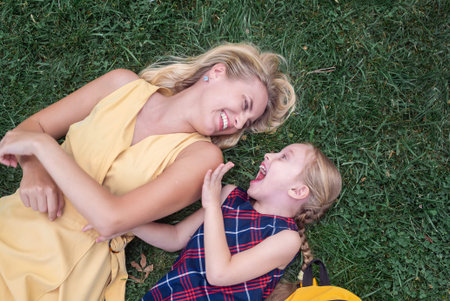 Mother and schoolgirl daughter lie on green grass in park laughing happily young woman and girl talk resting after school child shares impressions of lessons smiling cheerfully upper viewの写真素材