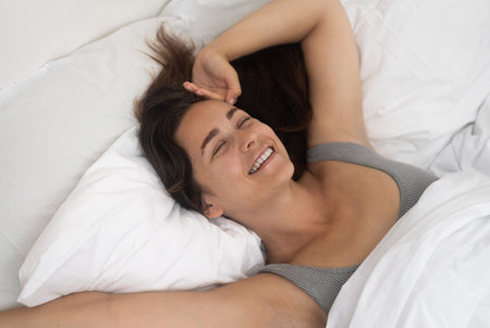 Smiling woman holds hand on forehead resting on bed with soft blanket in accommodation bedroom happy...