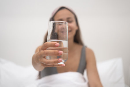 Smiling young woman holds out glass of water sitting on bed with light sleeping linen in bedroom of accommodation taking water after receiving energy from sleepの写真素材