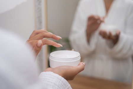 Woman hands holding lotion jar at dressing table in bathroom lady in gown taking anti aging skincare product from plastic bottle for applying in bathroom closeupの写真素材
