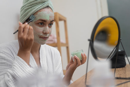 Woman applying homemade clay mask on face with brush sitting at table mirror lady in bathrobe using craft organic cosmetics to cleanse facial skin at homeの写真素材
