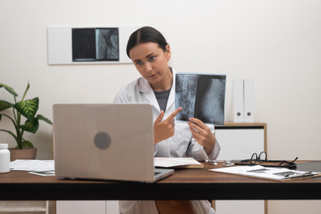 Virtual Medical Discussion: During a telemedicine video call, a doctor presents X-ray films on a laptop screen and elucidates the diseases characteristics to the patient.の写真素材