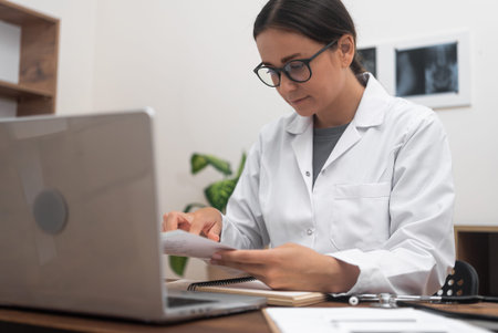 Lady doctor in glasses reads results of patient tests working at desk with laptop and documents...