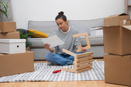 woman, sitting on the floor of her new house, assembles a self-assembly shelf amidst moving carton boxes and furniture, settling into her rental homeの写真素材