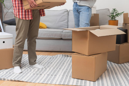 couple stands in their new homes living room, surrounded by a stack of cardboard boxes from their...