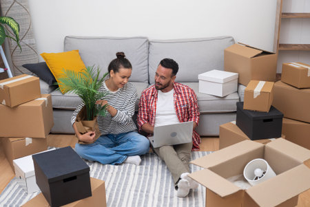 Starting Anew: Couple, homeowners or renters, on moving day, sit among boxes, using a laptop to select interior designs, shop online, or organize their move.の写真素材