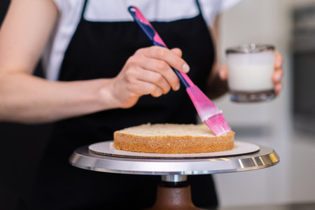 Skilled female confectioner smearing gluten free cake layers with fresh cream using pink brush woman in black apron preparing delicious confectionery from organic products closeupの写真素材