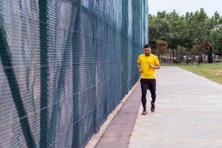 Engaged in activities to improve health and body, a bearded man in a tracksuit runs along a road by a high mesh fence on a sports ground in an urban park.の写真素材