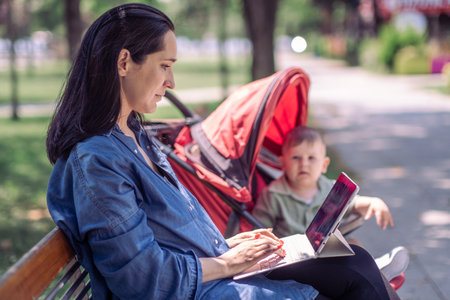 Concentrated woman using laptop for work tasks while baby son trying to get out of carriage in shady summer park freelancer and toddler needing attentionの写真素材