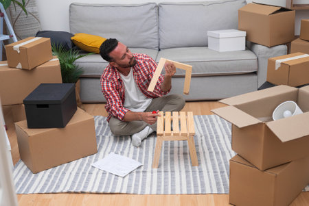 Brunette man with beard tries to figure out correct assembly of wooden chair sitting with serious expression on floor near sofa among cardboard boxes in spacious premiseの写真素材