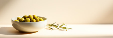 Bowl of bright green olives under soft light with olive leaves on a clean, neutral background.の素材