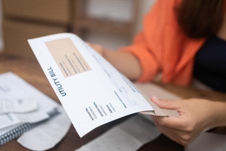 serious woman scrutinizes utility statements, tallying up expenses as prices for electricity, water,...